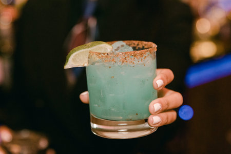 A bartender presents a colorful blue cocktail garnished with a lime slice and salt rim, showcasing...