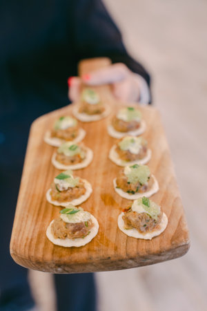 A server presents a wooden platter filled with elegant mini appetizers topped with greens at an evening gathering.の写真素材