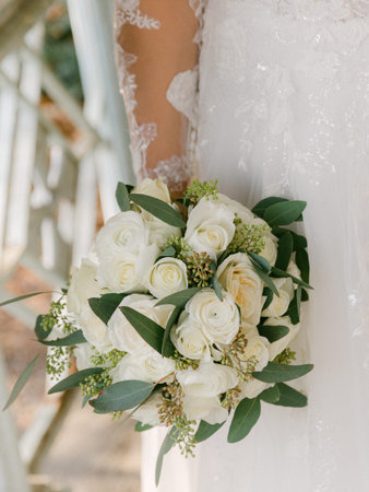 A bride in a lace dress gracefully holds a bouquet of white roses while standing amidst a tranquil outdoor venue.の写真素材