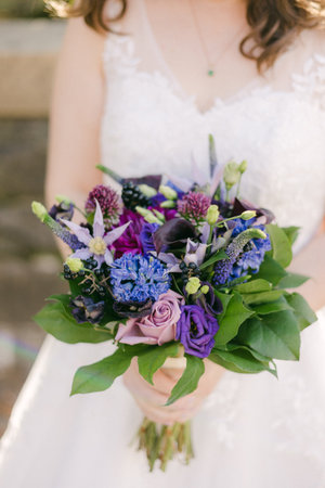 Bride displays a vibrant bouquet with purple and blue flowers while dressed in a wedding gown at an outdoor venue.の写真素材