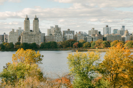 Colorful autumn trees frame the peaceful lake reflecting city skyscrapers under a cloudy sky.の写真素材