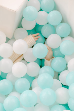 Two hands are visible reaching out from under a colorful pile of aqua and white balls in a playful...