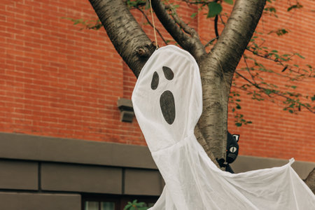A white ghost decoration dangles from a tree branch in a urban neighborhood, signaling the festive Halloween spirit.の写真素材
