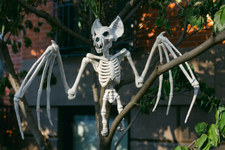 A spooky bat skeleton with white bones is dangling from a tree branch in a neighborhood, catching...
