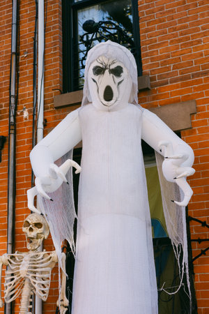 A tall ghost decoration stands beside a skeleton in front of a brick building during Halloween...