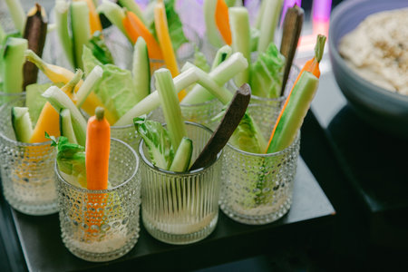 Fresh vegetable sticks served in elegant glasses beside a bowl of dip at an indoor eventの写真素材