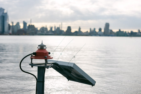 Solar panel with antennas sits by calm water, with a city skyline visible under a cloudy sky in the distance.の写真素材