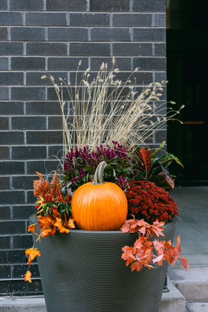 Bright orange pumpkin surrounded by vibrant autumn flowers and grasses in a stylish pot against a brick backdrop.の写真素材