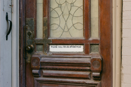 An ornate wooden door with a glass panel and a sign asking visitors to call for an appointment in a calm setting.の写真素材