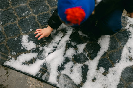 A young child explores the snow-covered welcome mat, making patterns with their hands on a cold...