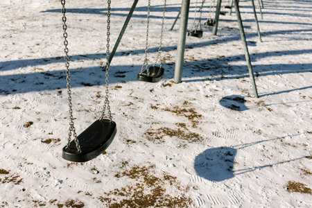 Empty swings sway gently in a snowy playground, creating shadows on the frosty ground as the sun...