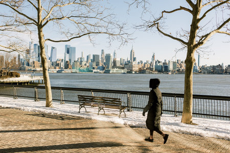 A person strolls along a snowy path by the water, enjoying the view of the skyline under clear skies.の写真素材