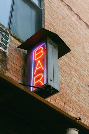 A bright neon 'BAR' sign shines against a brick building on a lively street during the evening...