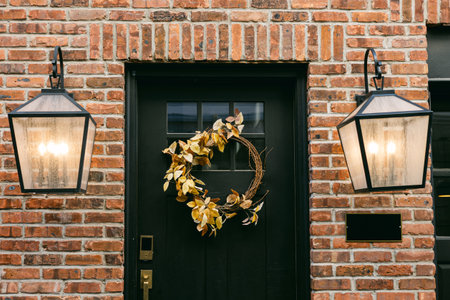 A cozy entryway with a black door, fall wreath, and glowing lanterns against a brick wall.の写真素材
