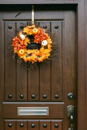 A vibrant autumn wreath, filled with orange flowers and pumpkins, decorates a wooden door, welcoming...