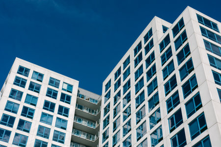 Two modern apartment buildings with large windows stand against a bright blue sky, symbolizing city...