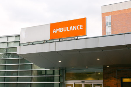 An orange sign displaying the word ambulance hangs above the entrance to a hospital building on a cloudy day.の写真素材