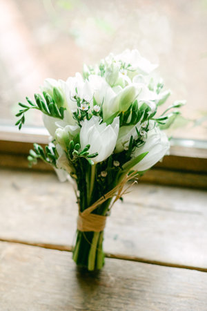 A charming bouquet of white flowers and greenery rests on a wooden table beside a window, illuminated by natural light.の写真素材