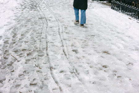 Footprints and tire tracks mark a snowy walkway as a person walks in winter weather in a local park.
