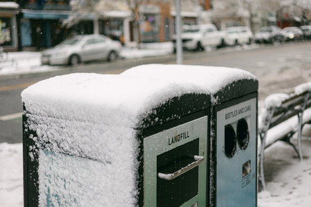 Snow covers recycling and landfill bins in a city street during winter weatherの写真素材