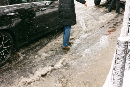 Person walks on icy street beside parked car during winter snowfall in urban settingの写真素材
