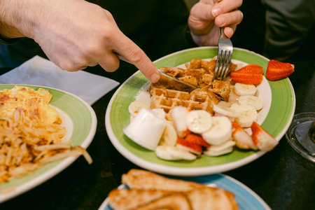 A person cuts a waffle while enjoying a plate with fruit and breakfast items at a table in a...