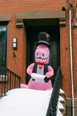 A pink dinosaur wearing a top hat and bow tie stands on the snowy steps of a brick house on a winter...