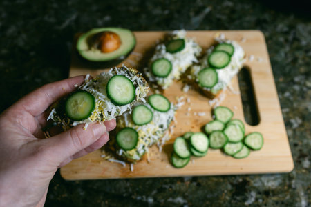 Cucumber slices are placed on a dish with avocado. A hand holds a piece above the cutting board.