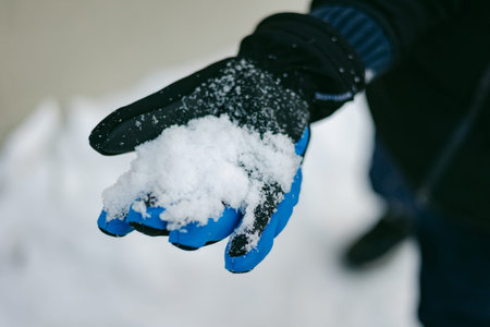A person holds a handful of snow with a blue glove in a snowy setting on a winter day.