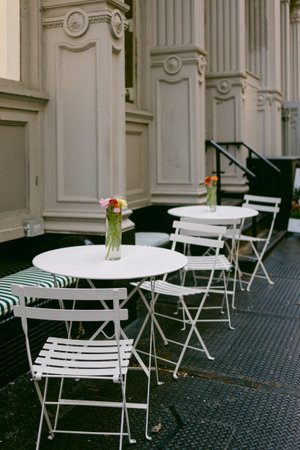 Tables and chairs are arranged outside a building with flowers in vases, showing a city scene during...