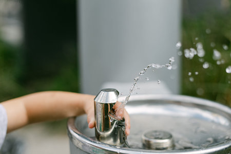 A child uses a water fountain in a park to drink water. The fountain has a metal spout and water sprays out. Other children can be seen playing nearby.の写真素材