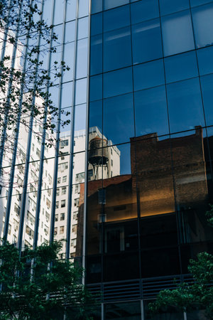 Several buildings reflect in the glass surface of a modern skyscraper. Tall trees are present in the...
