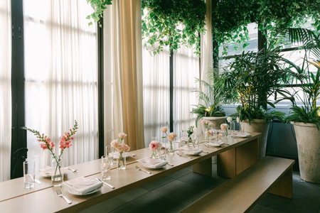 Tables are set for dining in a restaurant space with green plants and flowers. Sunlight shines...