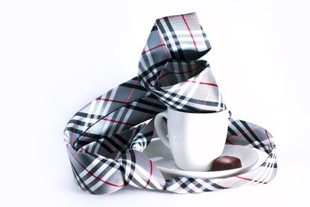 Checkered necktie lie on the table twisted around a cup of coffee  A little chocolate candy lie in the white ceramic saucer near ceramic cup  Objects are isolated; background is white の写真素材