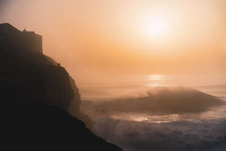 Silhouette of a man sitting on a cliff watching a giant wave breaking with the sunset in the background.の写真素材