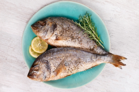 Two grilled barbeque sea bream on plate with fresh rosemary herbs and lemon on white wooden...
