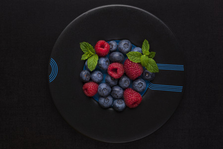 Fresh berries on black plate on black background, top view. Raspberries and blueberries with fresh mint leaf, minimal modern style.の写真素材
