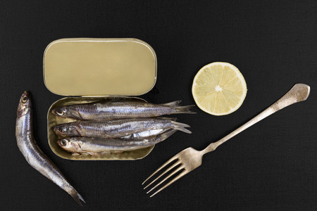 Fresh anchovy fish in can with silver fork isolated on black textured background, top view. Culinary healthy seafood eating.の写真素材
