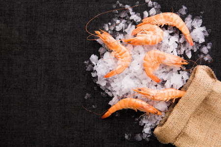 Fresh shrimp with lemon on ice in brown burlap bag on black table, top view. Culinary seafood eating.の写真素材