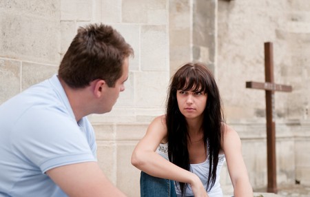 Young couple sitting outdoors ahead of church (cross in background) having relationship problemsの写真素材