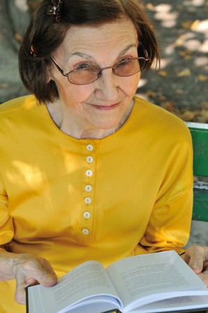 Senior woman sitting on bench reading book outdoors in parkの写真素材