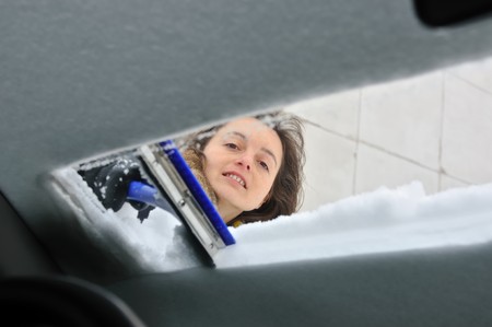 Young smiling woman cleaning car windows from snow in winter season