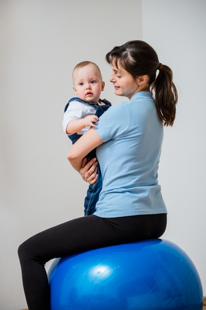 Mother exercises with blue fit ball at home holding her baby on handsの写真素材