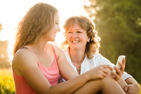 Teenage girl showing her mother photos on mobile phone outdoor in nature with setting sun in backgroundの写真素材
