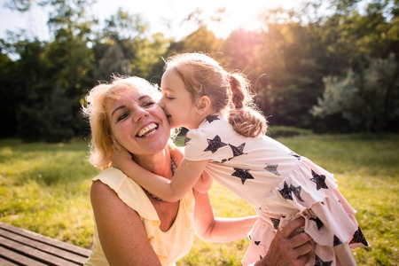 Grandchild kissing her happy grandmother on cheek - outdoor in nature with sun in backgroundの写真素材