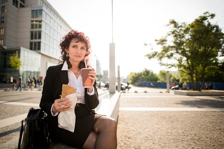Senior business woman on fast food lunch - drinking take-away coffee and eating sandwich in streetの写真素材