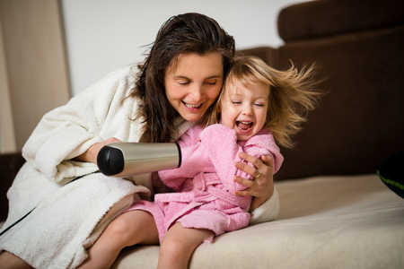 Mother and her little daughter in bathrobes having fun with hairdryerの写真素材