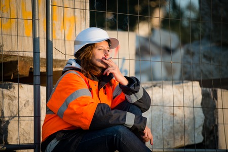 Senior woman engineer in protective workwear relaxing during break and smoking cigarette
