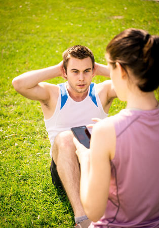 Man making sit-ups while woman is watching time of exercise on mobile phone