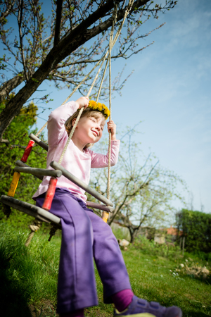 Little girl with dandelion wreath swnging on seesaw hanging from tree in backyardの写真素材